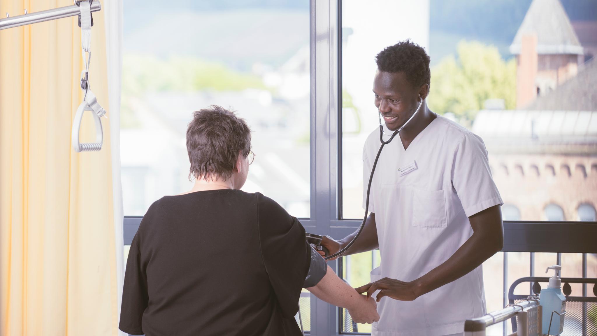 Man sieht einen Mann im Pflegergewand der einer Patientin, die auf einem Bett sitzt, den Puls misst