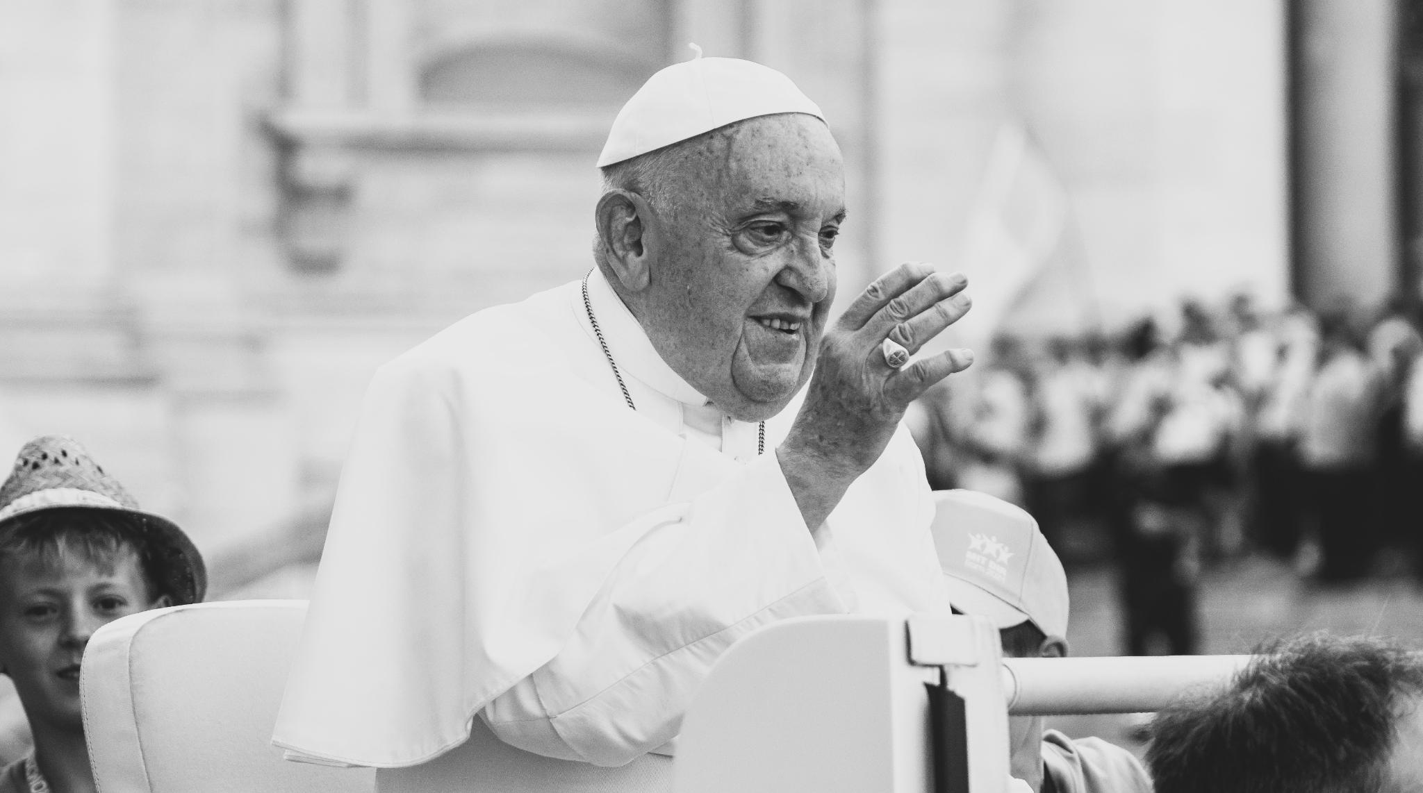 Schwarzweiß-Foto zeigt Papst Franziskus, wie er winkend im Papamobil an Menschen vorbeifährt.