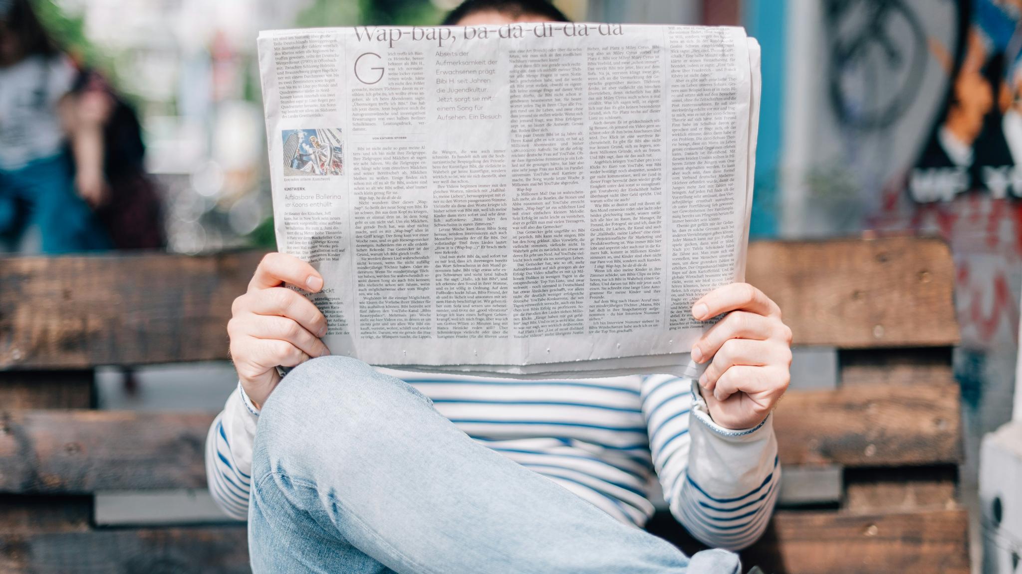 Man sitzt auf einer Bank und hät eine großformatige Zeitung in Händen, die sein Gesicht verdeckt.