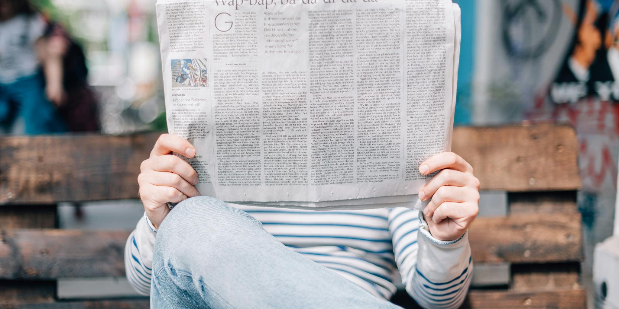 Man sitzt auf einer Bank und hät eine großformatige Zeitung in Händen, die sein Gesicht verdeckt.