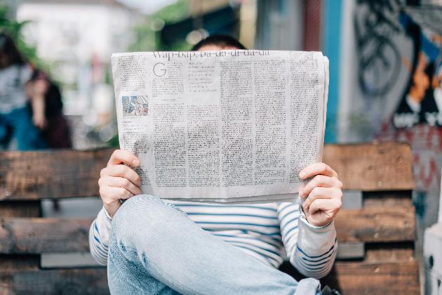 Man sitzt auf einer Bank und hät eine großformatige Zeitung in Händen, die sein Gesicht verdeckt.