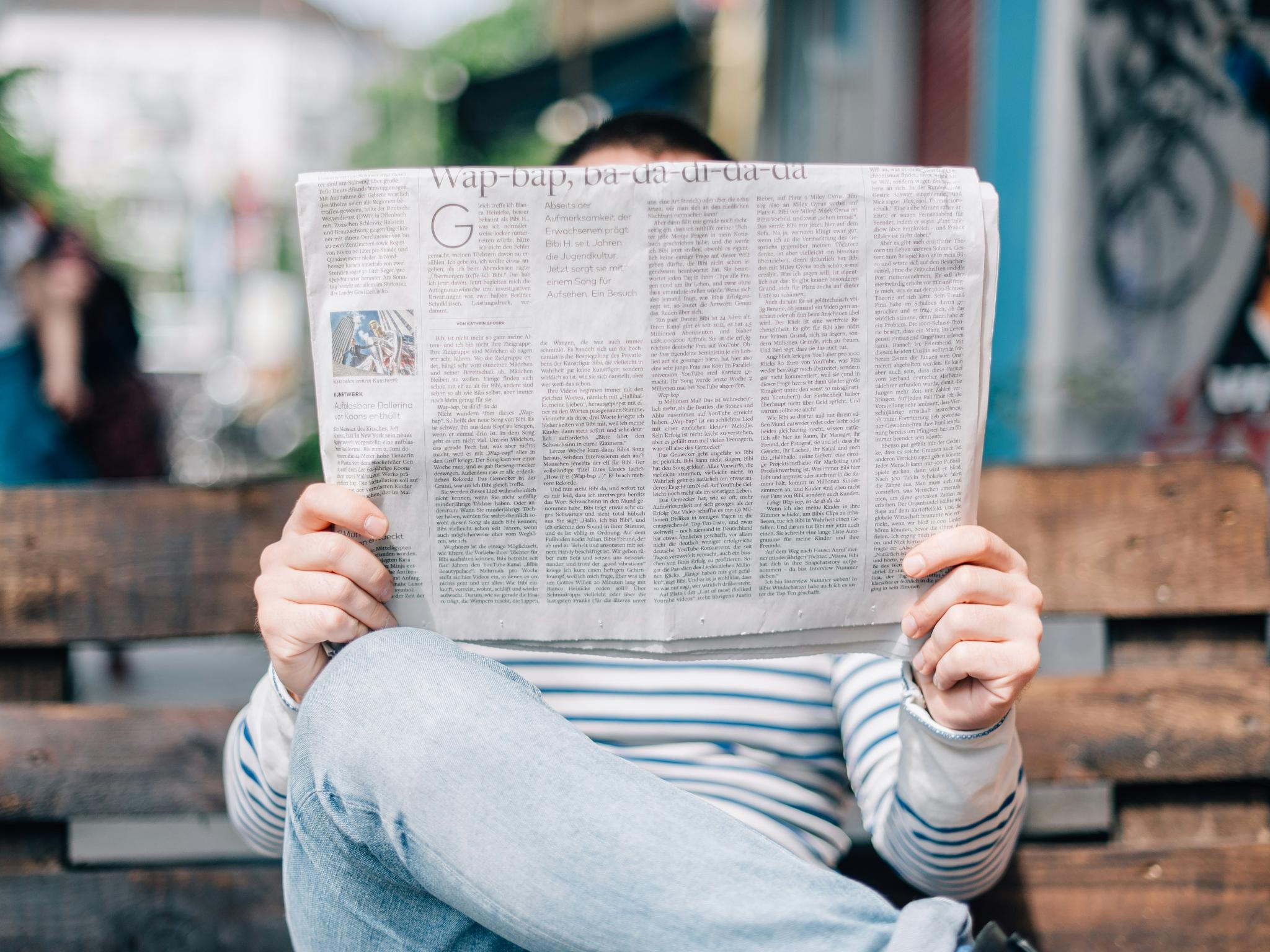 Man sitzt auf einer Bank und hät eine großformatige Zeitung in Händen, die sein Gesicht verdeckt.