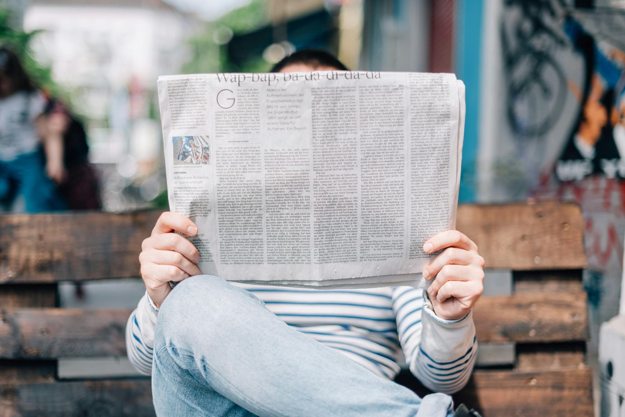 Man sitzt auf einer Bank und hät eine großformatige Zeitung in Händen, die sein Gesicht verdeckt.