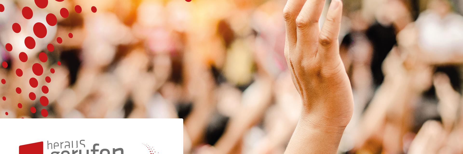 Eine Hand streckt sich in die Höhe, im Hintergrund sind verschwommen noch zahlreiche Menschen und hochgehobene Hände zu sehen.