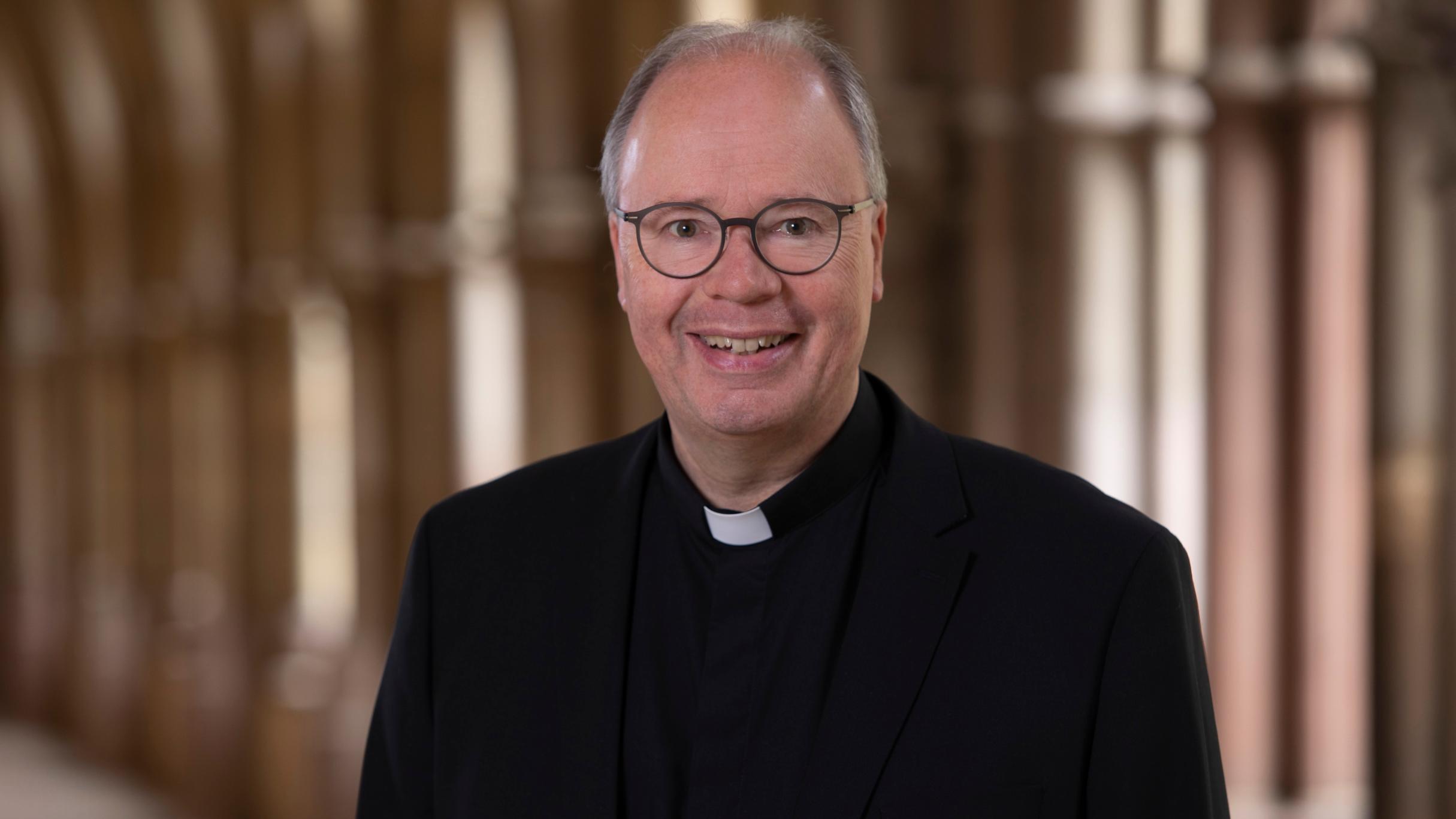 Man sieht Bischof Stephan Ackermann im Anzug im Dom Kreuzgang. Er lächelt in die Kamera