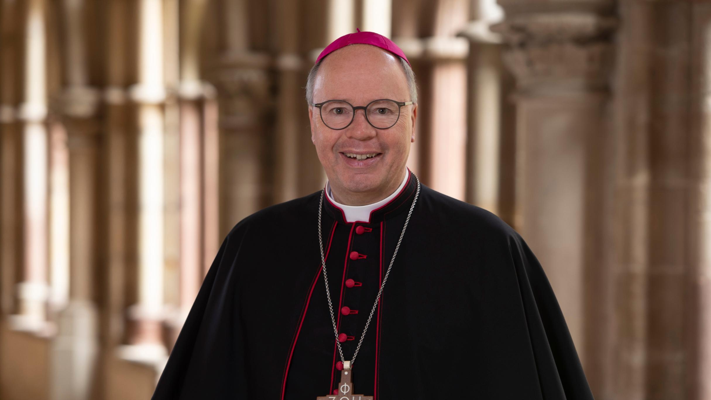 Man sieht Bischof Stephan Ackermann im Talar im Dom Kreuzgang. Er lächelt in die Kamera