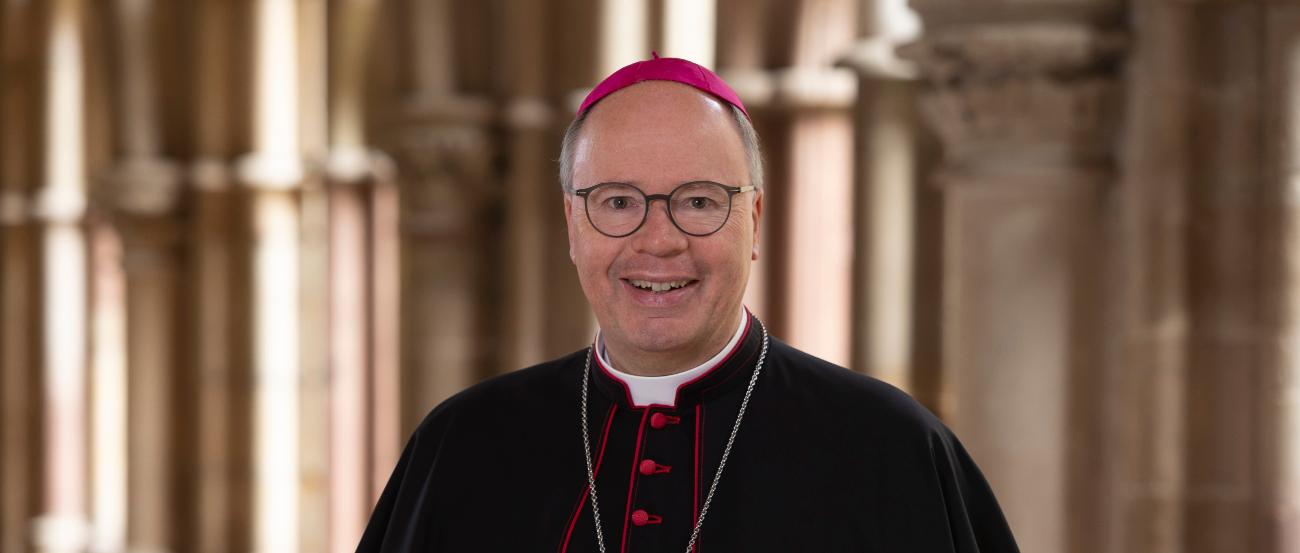 Man sieht Bischof Stephan Ackermann im Talar im Dom Kreuzgang. Er lächelt in die Kamera