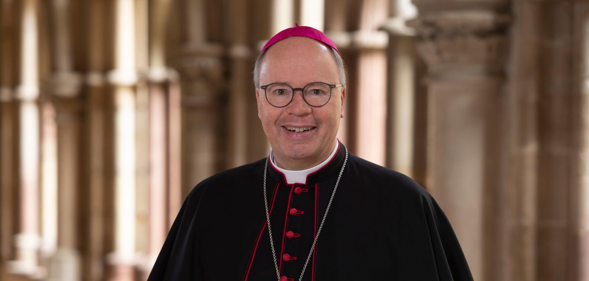 Man sieht Bischof Stephan Ackermann im Talar im Dom Kreuzgang. Er lächelt in die Kamera