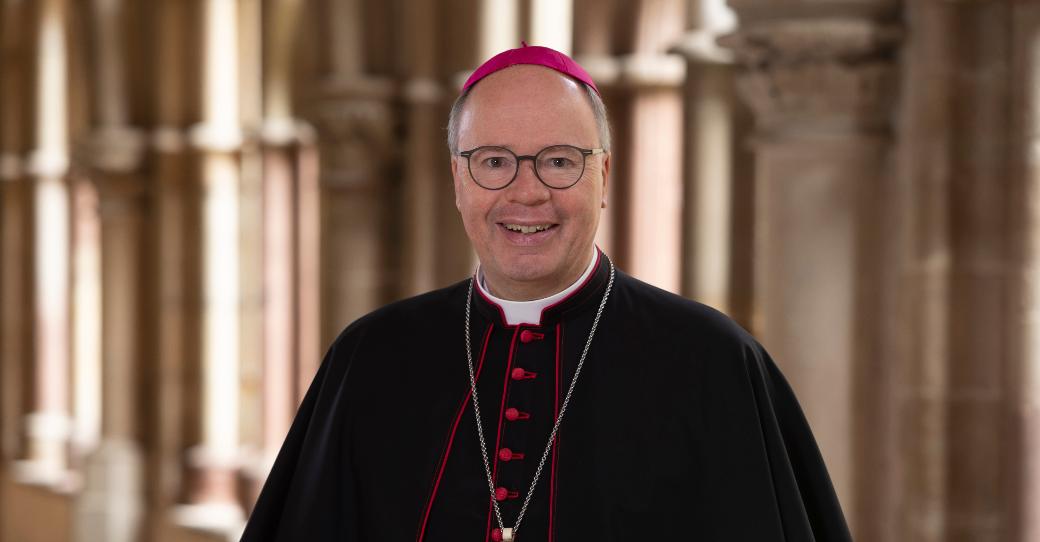 Man sieht Bischof Stephan Ackermann im Talar im Dom Kreuzgang. Er lächelt in die Kamera