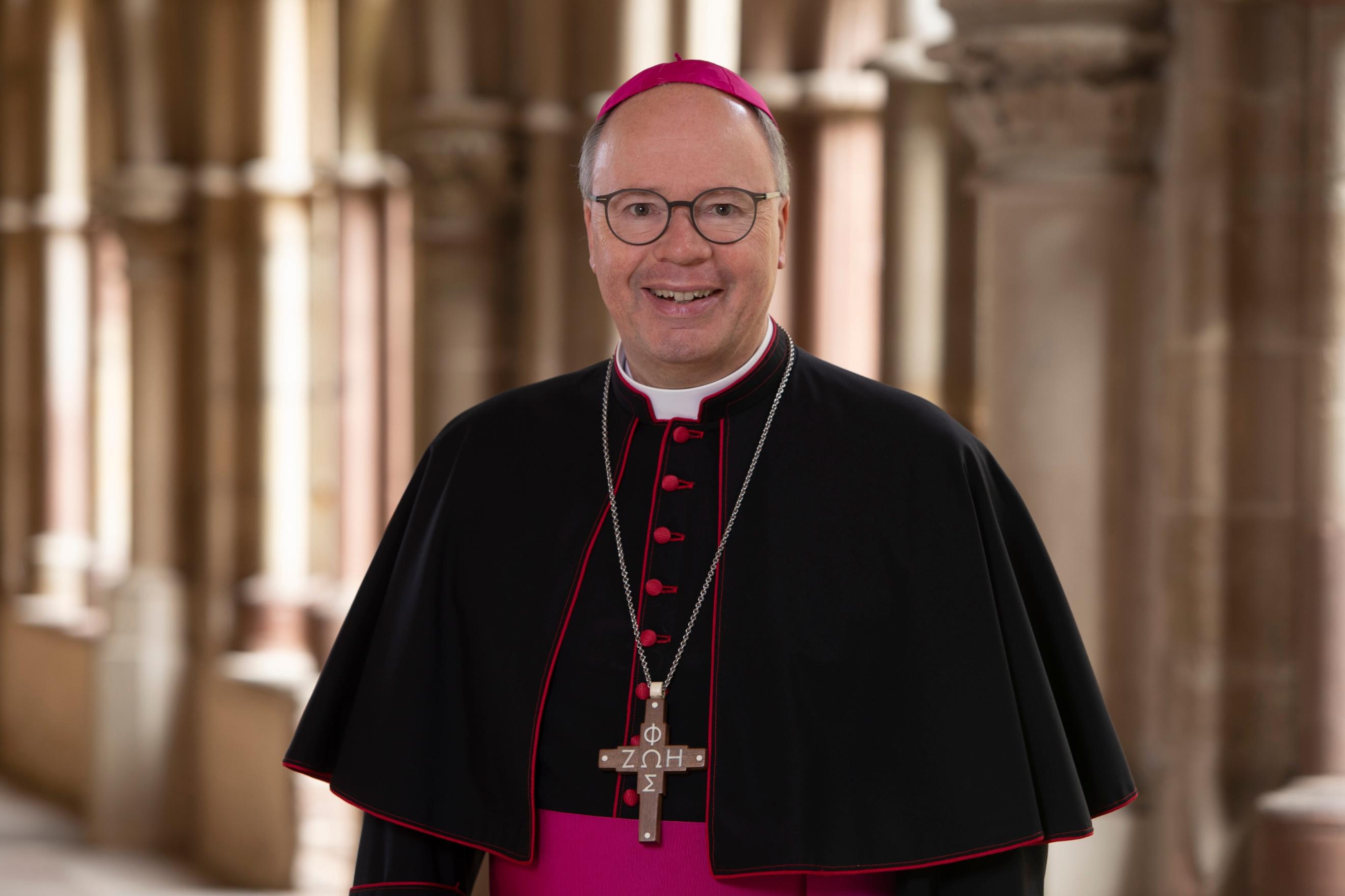 Man sieht Bischof Stephan Ackermann im Talar im Dom Kreuzgang. Er lächelt in die Kamera