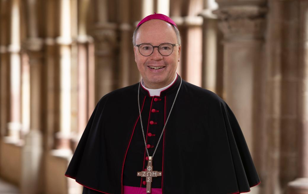 Man sieht Bischof Stephan Ackermann im Talar im Dom Kreuzgang. Er lächelt in die Kamera