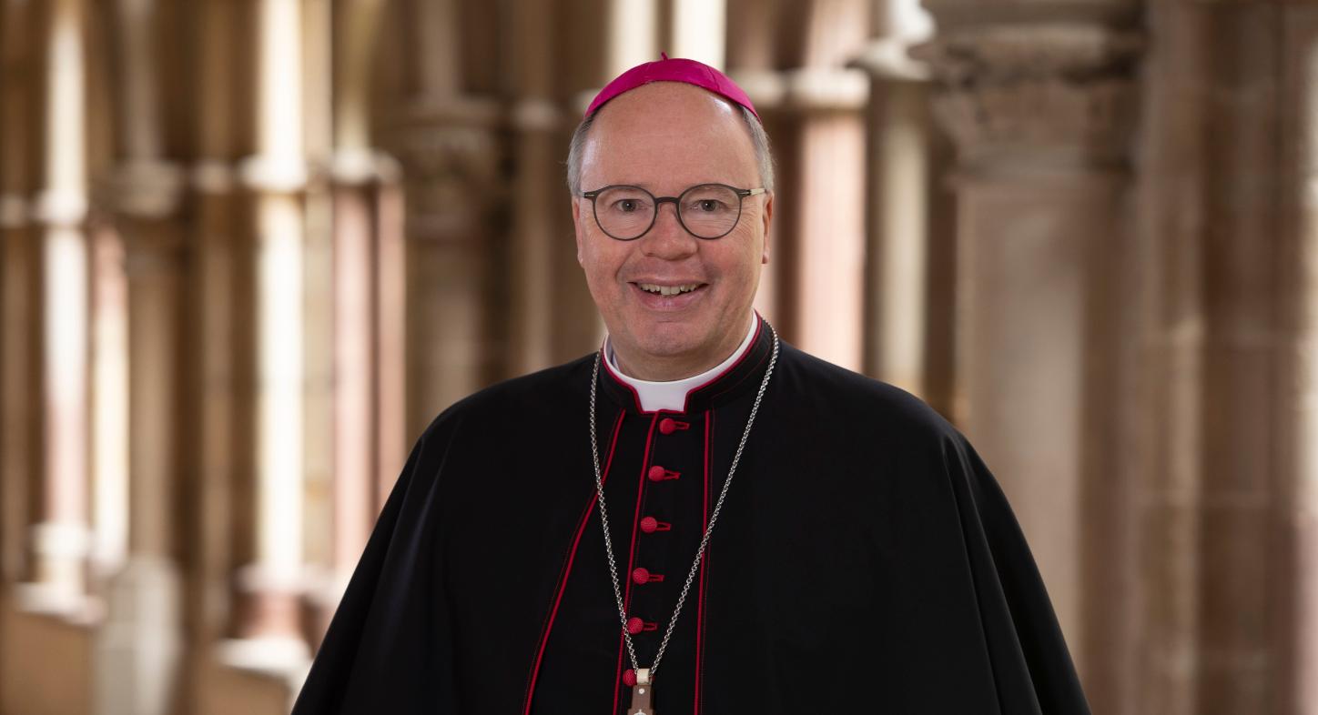 Man sieht Bischof Stephan Ackermann im Talar im Dom Kreuzgang. Er lächelt in die Kamera