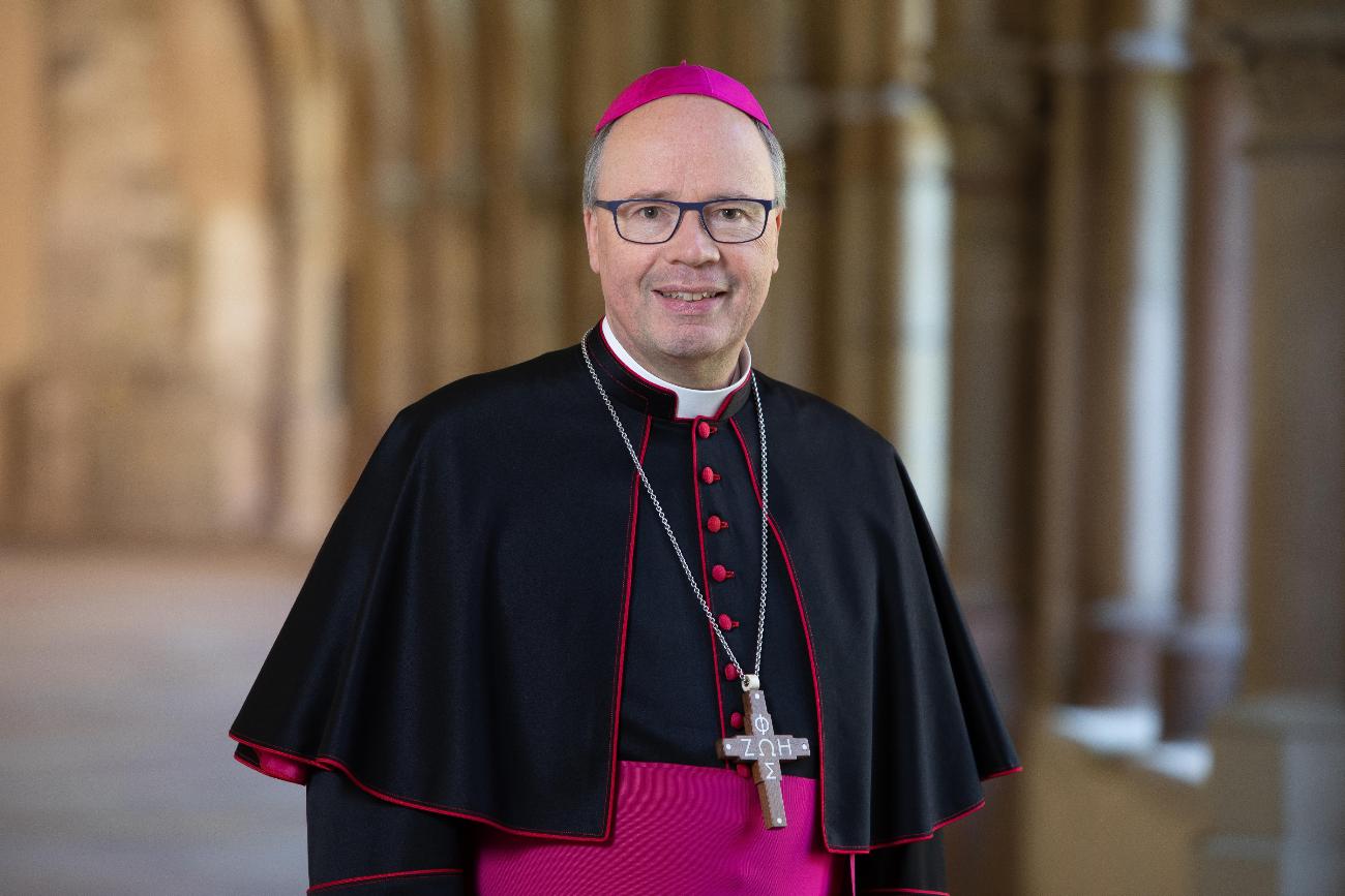 Hier ist ein Portrait von Bischof Ackermann, lächelnd im Kreuzgang zu sehen. Er Trägt ein Ornat.