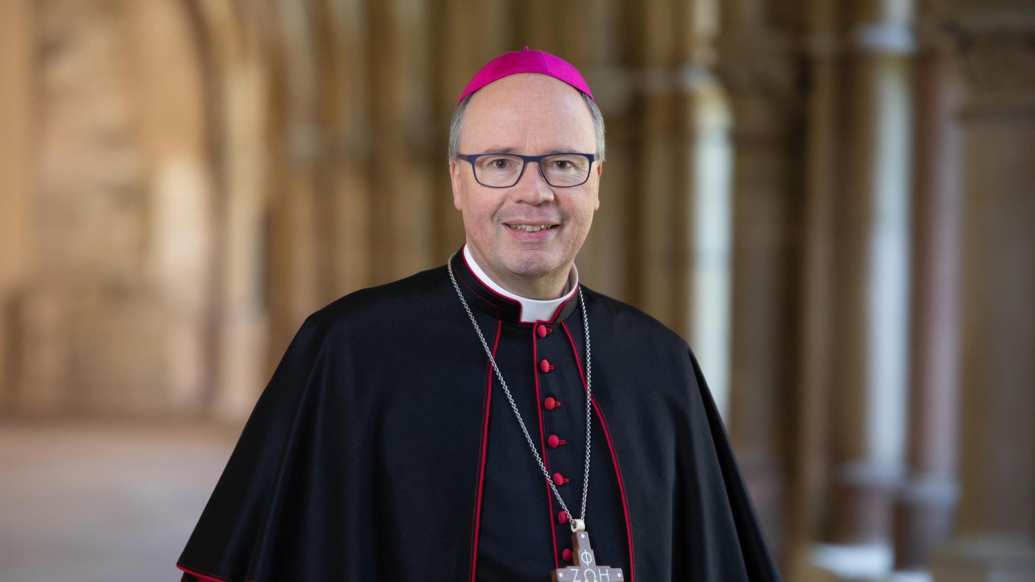 Hier ist ein Portrait von Bischof Ackermann, lächelnd im Kreuzgang zu sehen. Er Trägt ein Ornat.