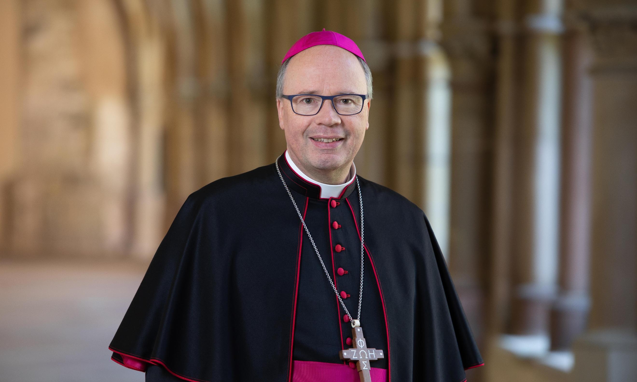 Hier ist ein Portrait von Bischof Ackermann, lächelnd im Kreuzgang zu sehen. Er Trägt ein Ornat.