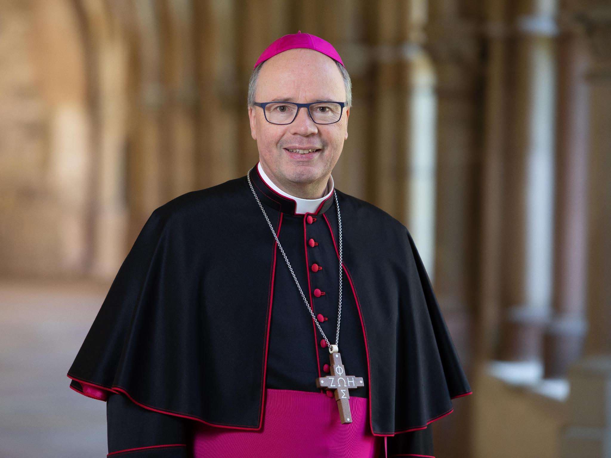 Hier ist ein Portrait von Bischof Ackermann, lächelnd im Kreuzgang zu sehen. Er Trägt ein Ornat.