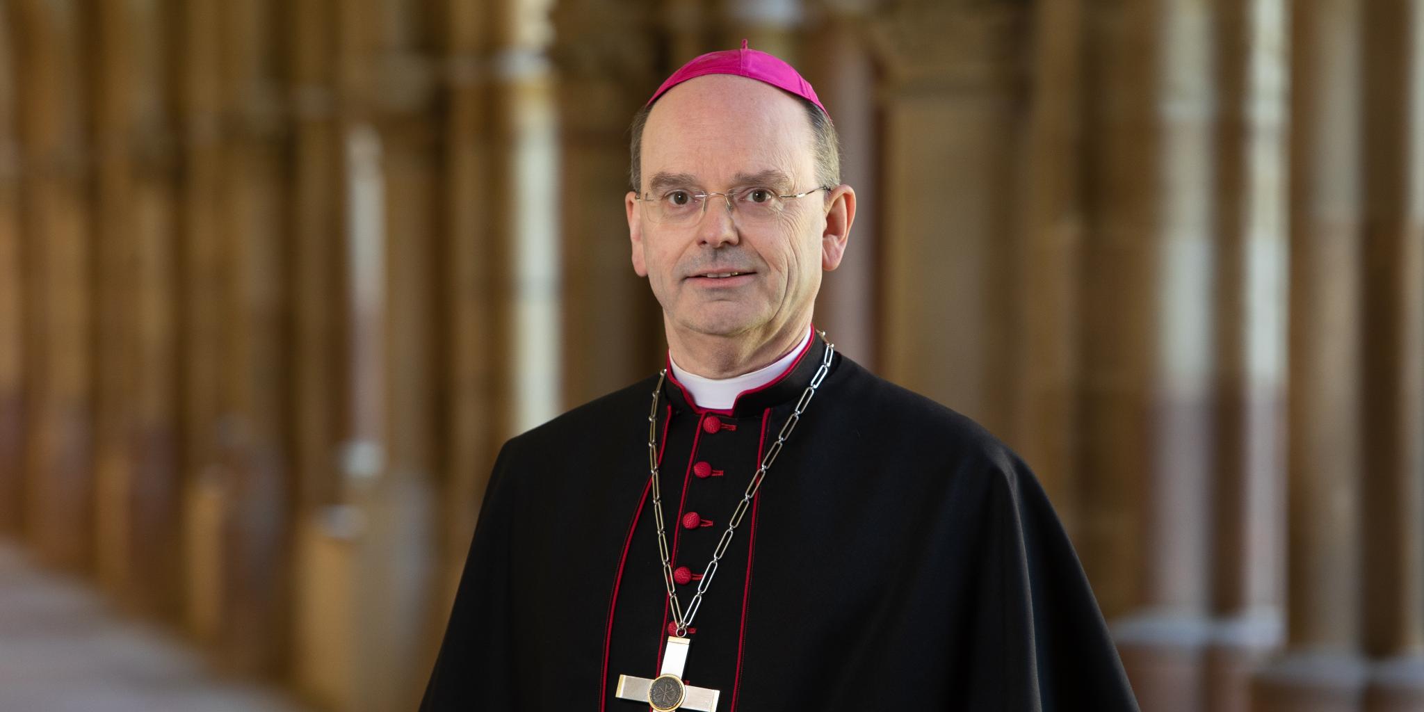 Hier ist Weihbischof Brahm lächelnd im Kreuzgang zusehen, zudem trägt er ein Ornat.