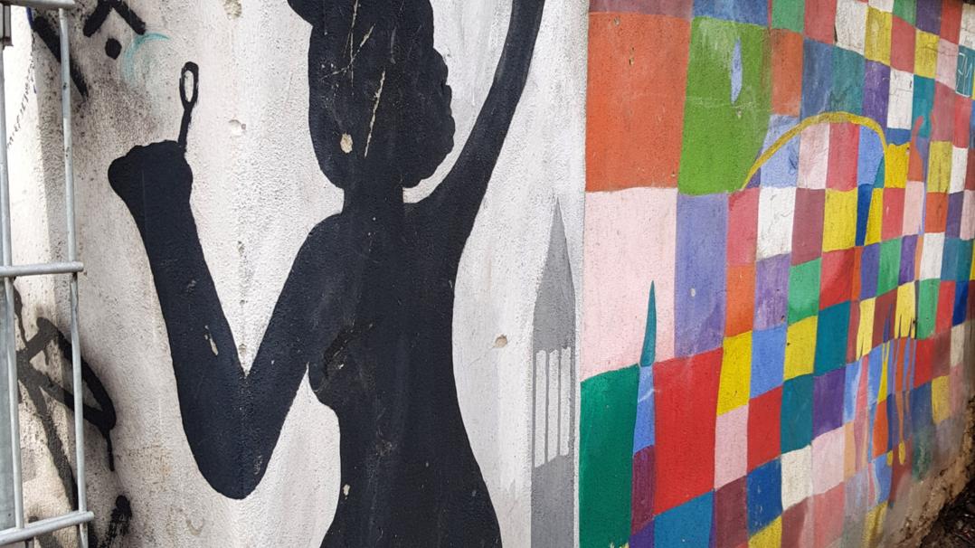 Graffiti an einer Wand. Man sieht den Schatten einer Frau mit Regenschirm. Ebenso viele bunte Kacheln