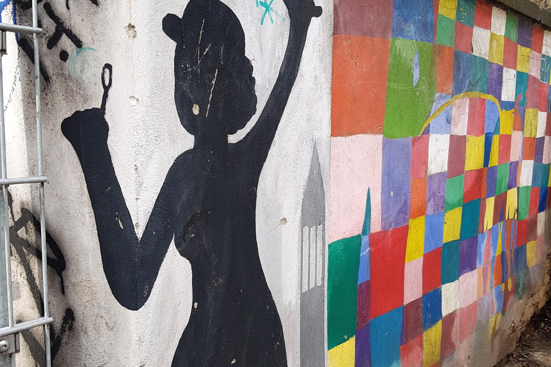 Graffiti an einer Wand. Man sieht den Schatten einer Frau mit Regenschirm. Ebenso viele bunte Kacheln