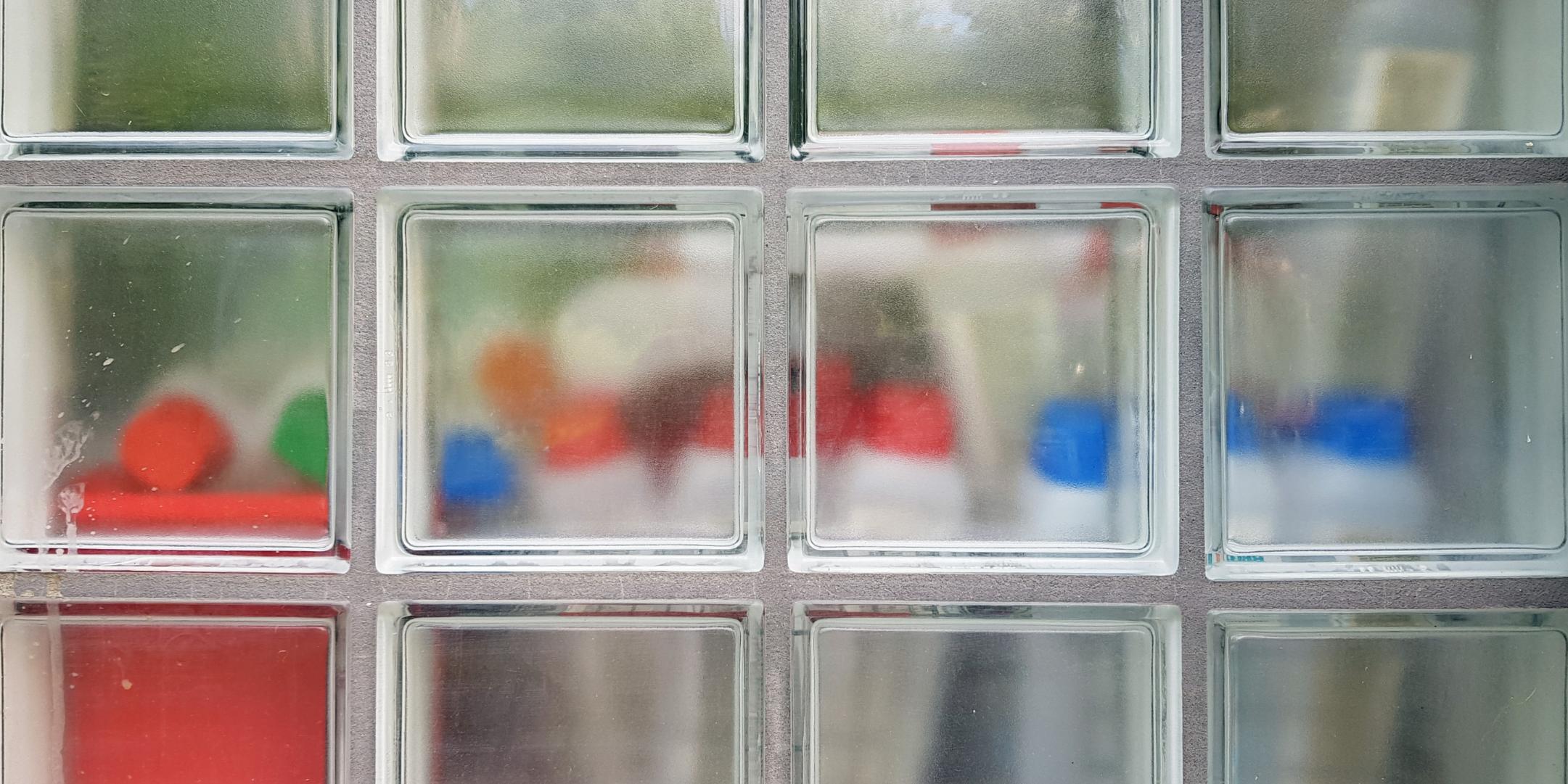 Foto einer Glasbausteinewand. Hinter den Glasbausteinen sind weiße Flaschen mit farbigen Köpfen zu sehen