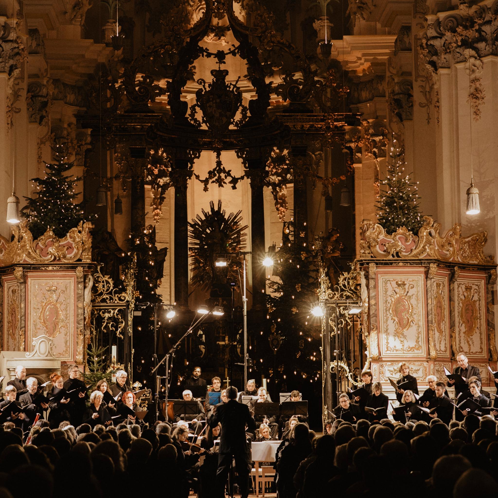 An einem Altar steht ein Chor, in schwarz gekleidet, die Kirche ist weihnachtlich dekoriert mit vielen Kerzen und Lichtern