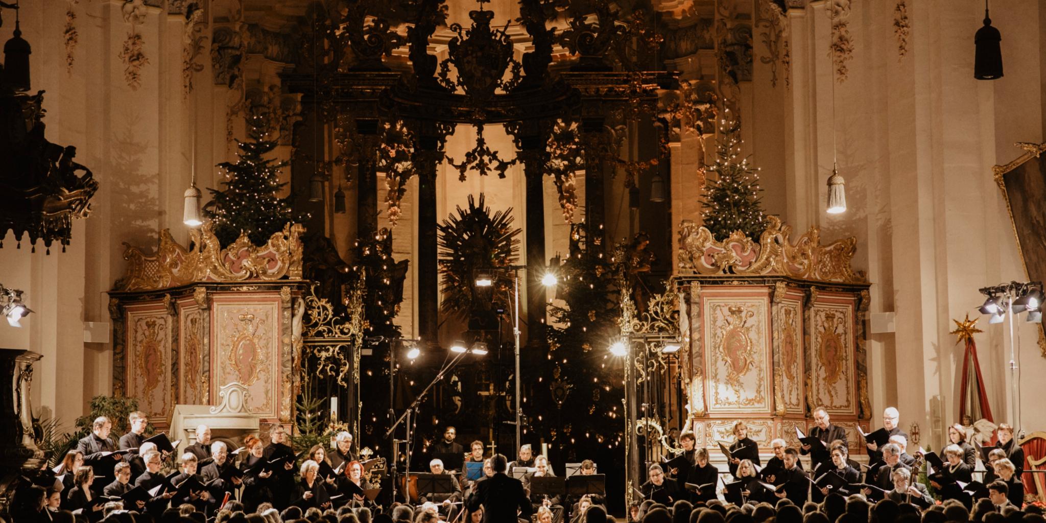 An einem Altar steht ein Chor, in schwarz gekleidet, die Kirche ist weihnachtlich dekoriert mit vielen Kerzen und Lichtern