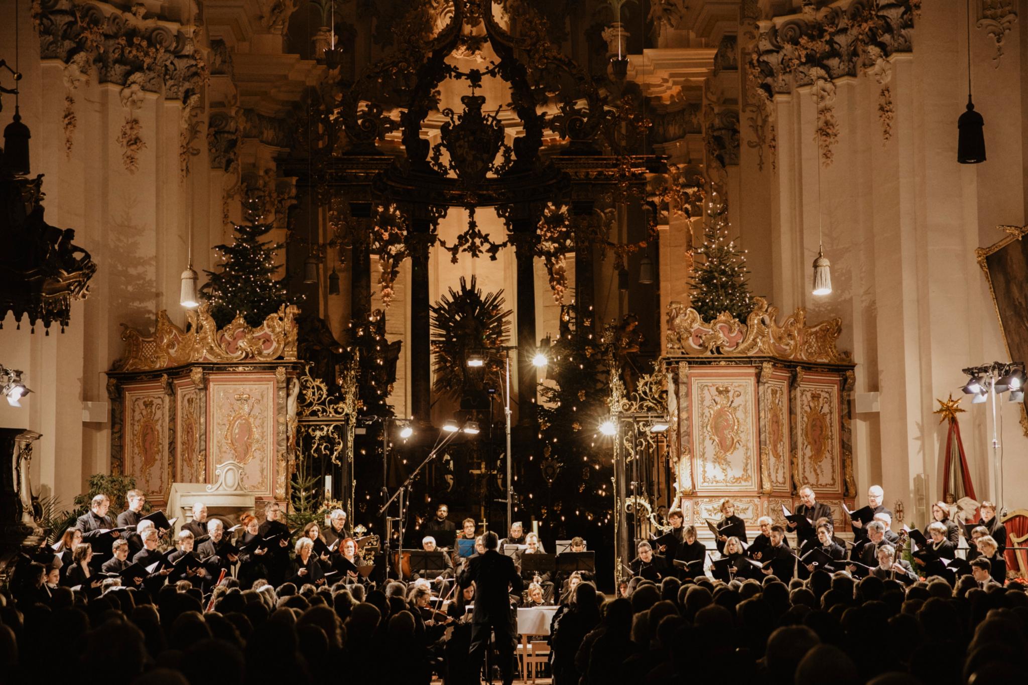 An einem Altar steht ein Chor, in schwarz gekleidet, die Kirche ist weihnachtlich dekoriert mit vielen Kerzen und Lichtern