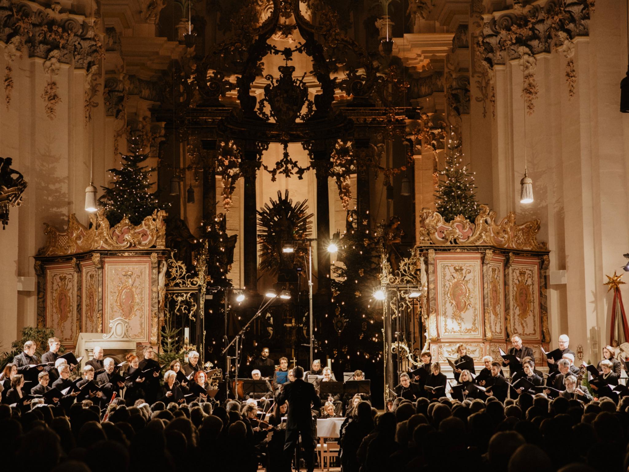 An einem Altar steht ein Chor, in schwarz gekleidet, die Kirche ist weihnachtlich dekoriert mit vielen Kerzen und Lichtern