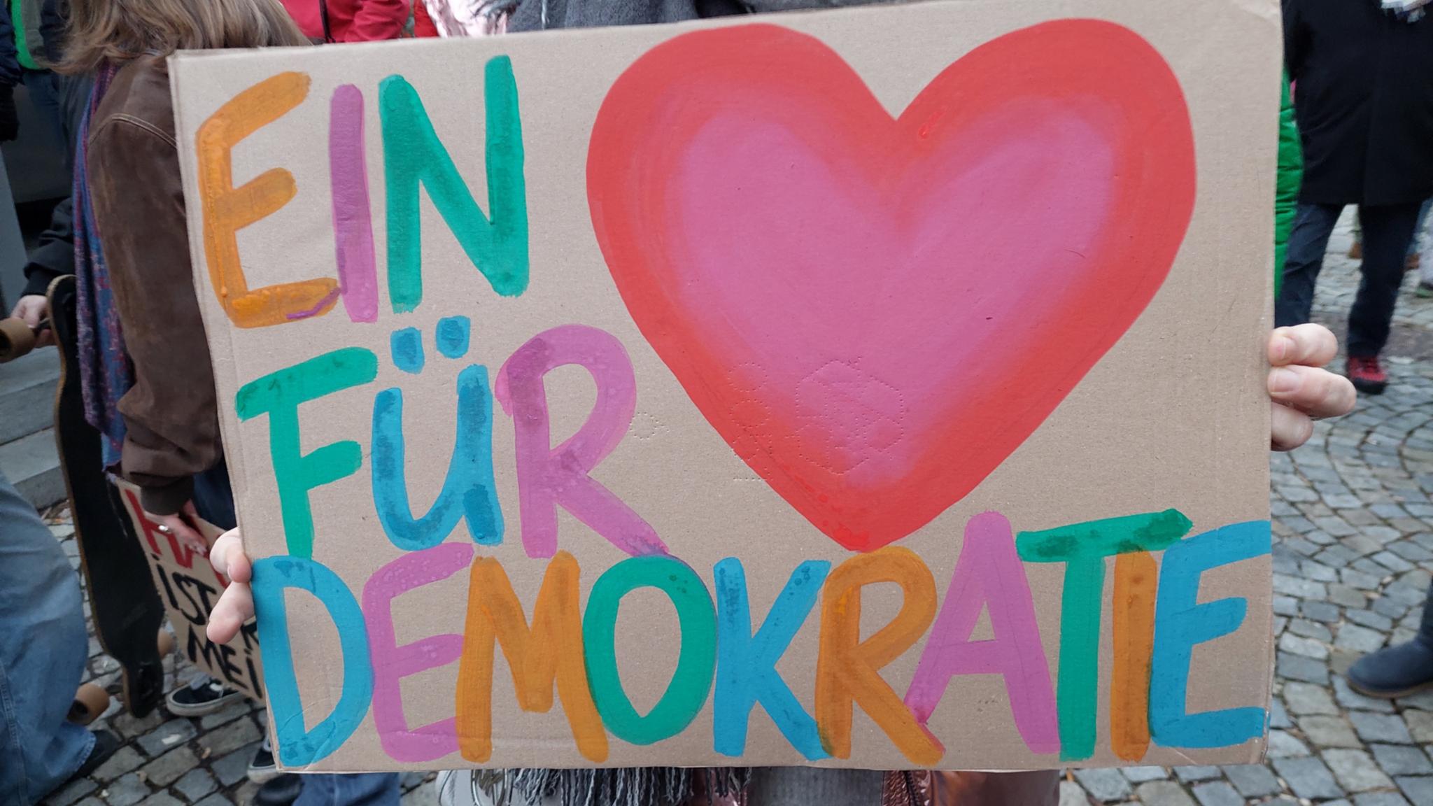 Auf einem Stück Pappe ist ein buntes Herz und in bunt steht ein Herz für Demokratie daneben.