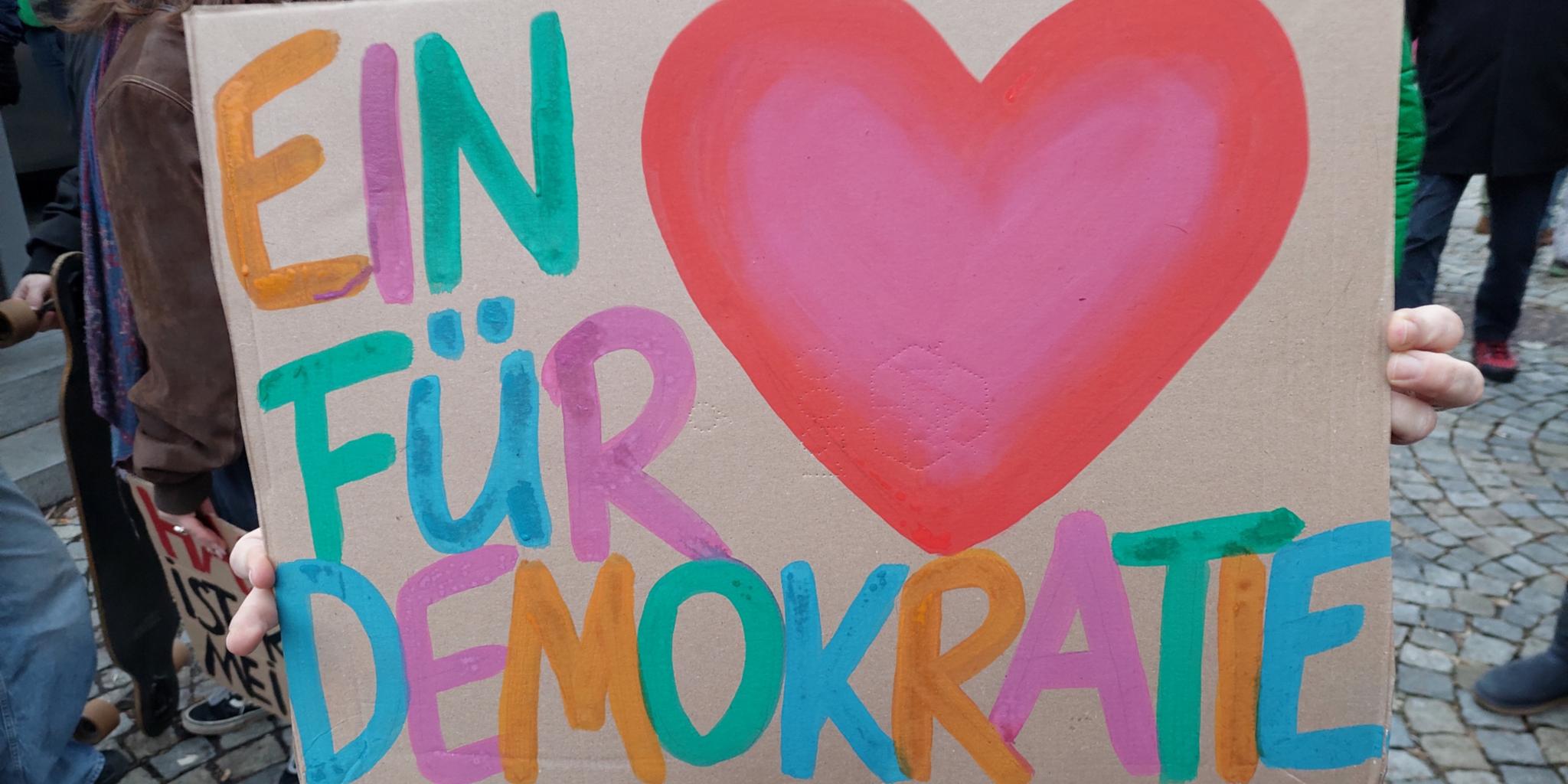 Auf einem Stück Pappe ist ein buntes Herz und in bunt steht ein Herz für Demokratie daneben.