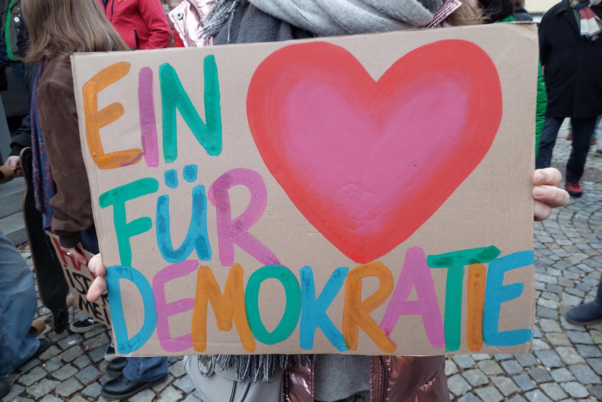 Auf einem Stück Pappe ist ein buntes Herz und in bunt steht ein Herz für Demokratie daneben.