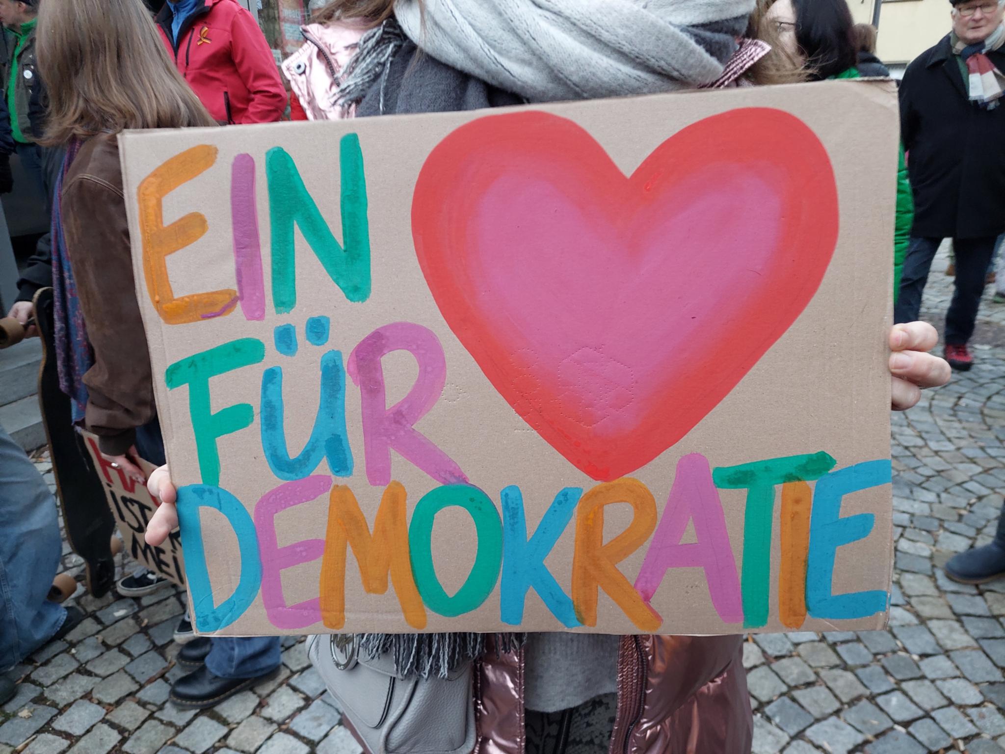 Auf einem Stück Pappe ist ein buntes Herz und in bunt steht ein Herz für Demokratie daneben.