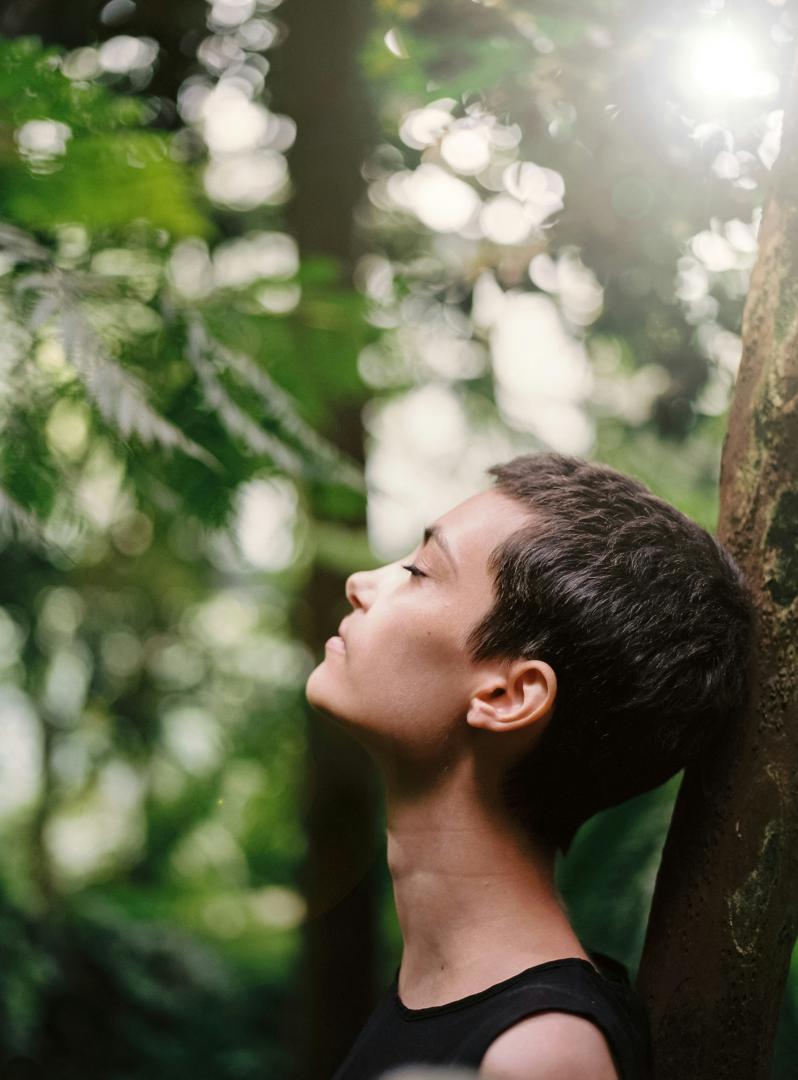 Eine Frau steht im Wald an einen Baum gelehnt und atmet tief ein