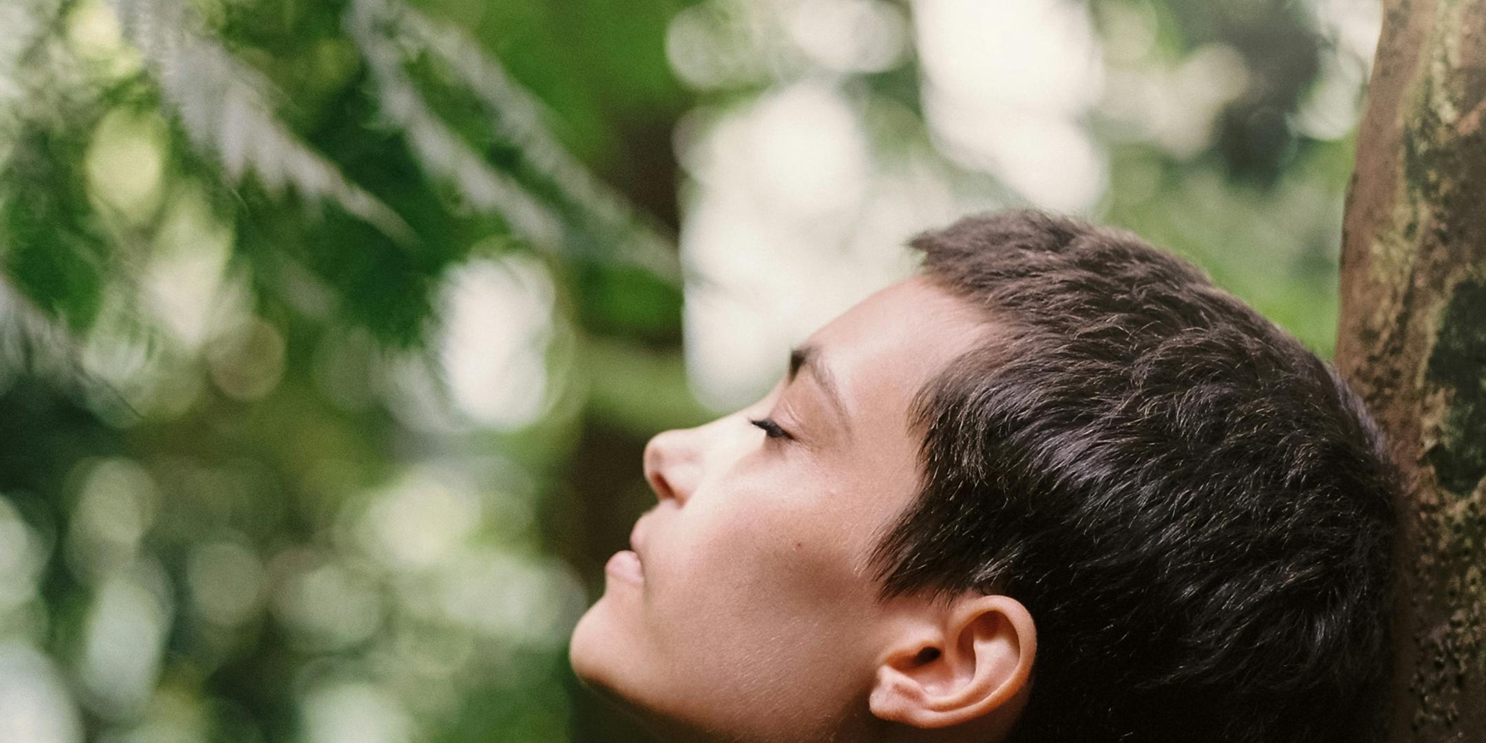 Eine Frau steht im Wald an einen Baum gelehnt und atmet tief ein