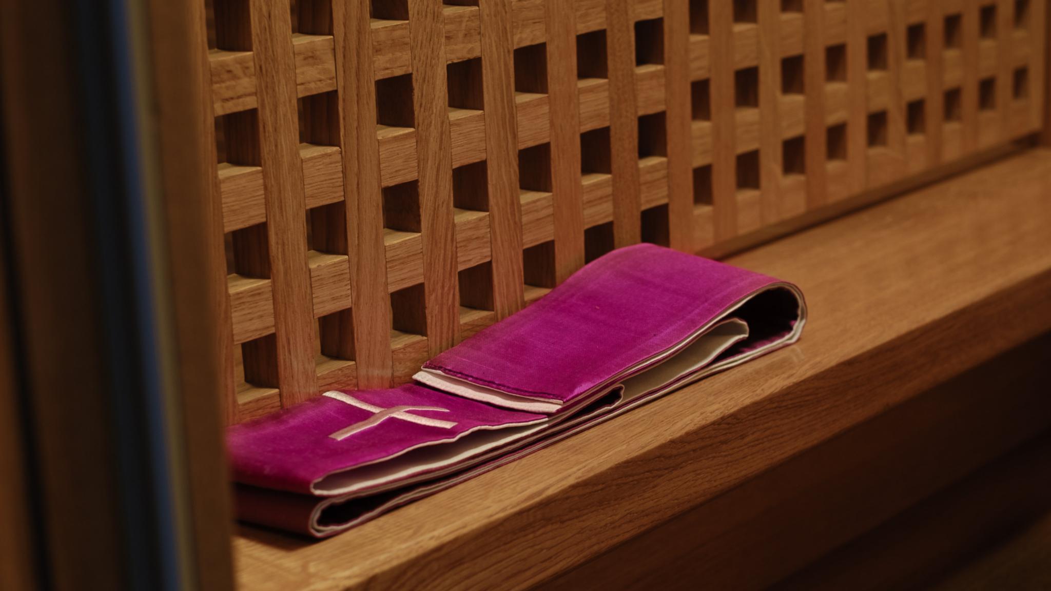 Man sieht als hölzernes Gitter eine Seite eines Beichstuhls. Auf der Bank davor liegt eine Priesterstola.