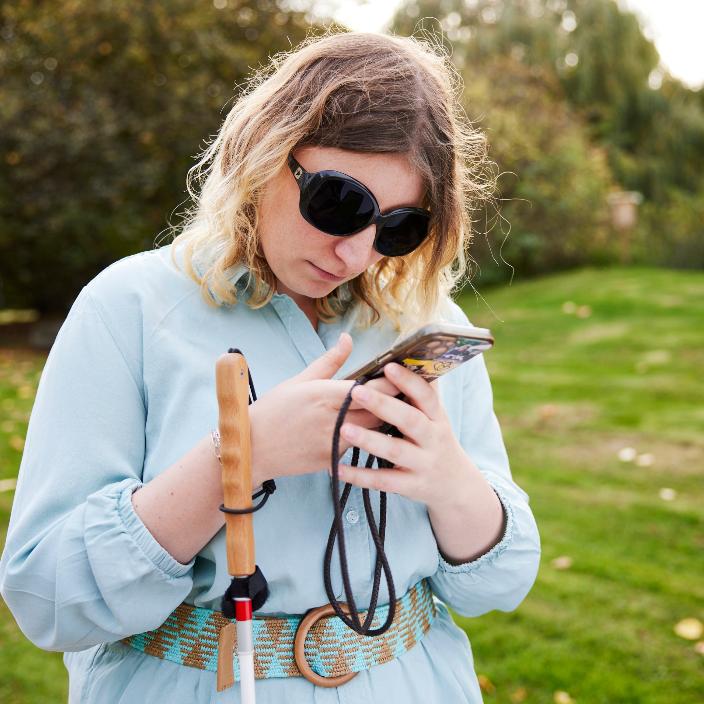 Eine blinde Frau mit Brille und Blindenstock steht in einem Park und hat ein Handy in den Händen und Kopfhörer in den Ohren.