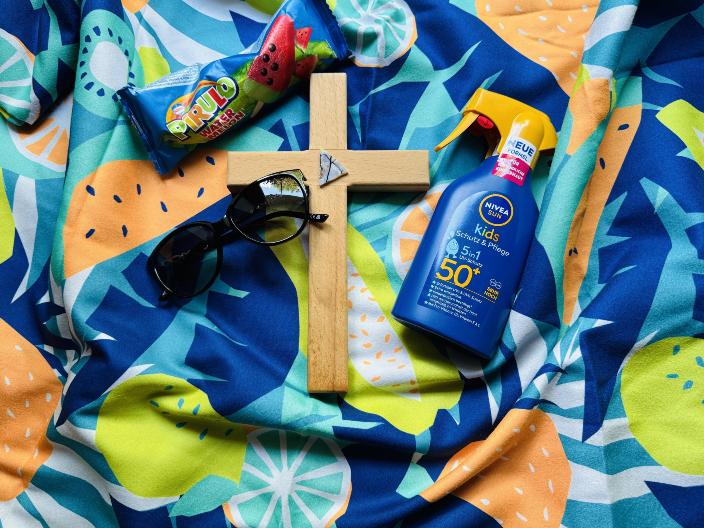 Auf einem bunten Badetuch liegt ein Kreuz, eine Sonnebrille, eine Flasche Sonnenmilch und ein Wassereis