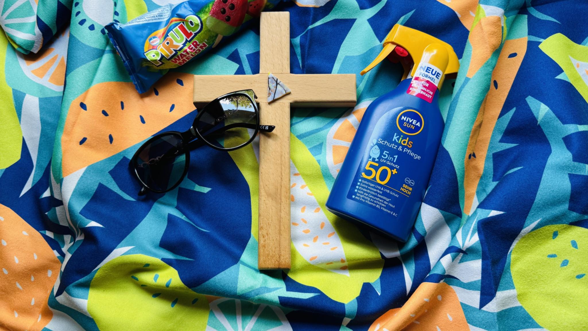 Auf einem bunten Badetuch liegt ein Kreuz, eine Sonnebrille, eine Flasche Sonnenmilch und ein Wassereis