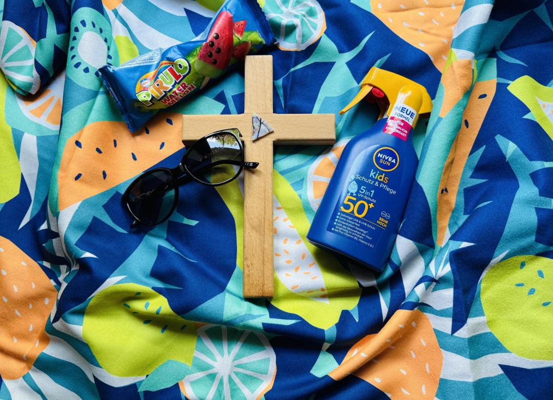 Auf einem bunten Badetuch liegt ein Kreuz, eine Sonnebrille, eine Flasche Sonnenmilch und ein Wassereis