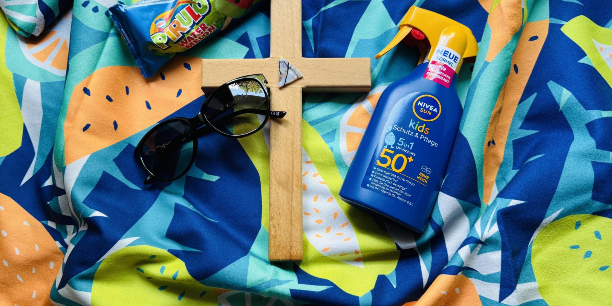 Auf einem bunten Badetuch liegt ein Kreuz, eine Sonnebrille, eine Flasche Sonnenmilch und ein Wassereis