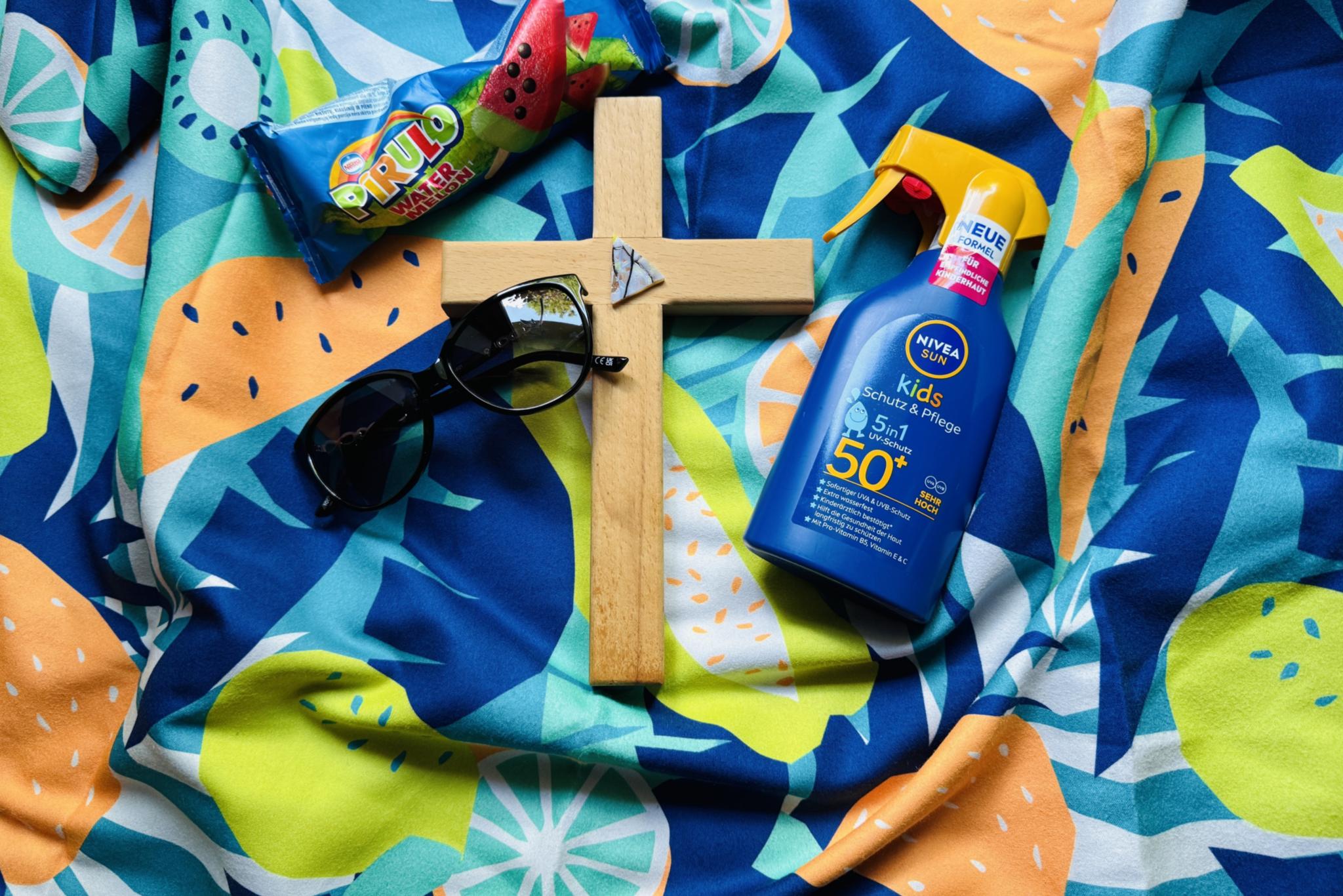 Auf einem bunten Badetuch liegt ein Kreuz, eine Sonnebrille, eine Flasche Sonnenmilch und ein Wassereis