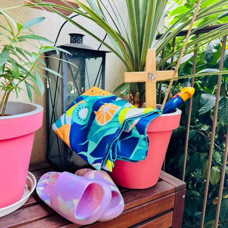 Zwischen Balkonpflanzen steht ein Kreuz, eine Flasche Sonnenmilch, Badeschuhe und ein Badetuch