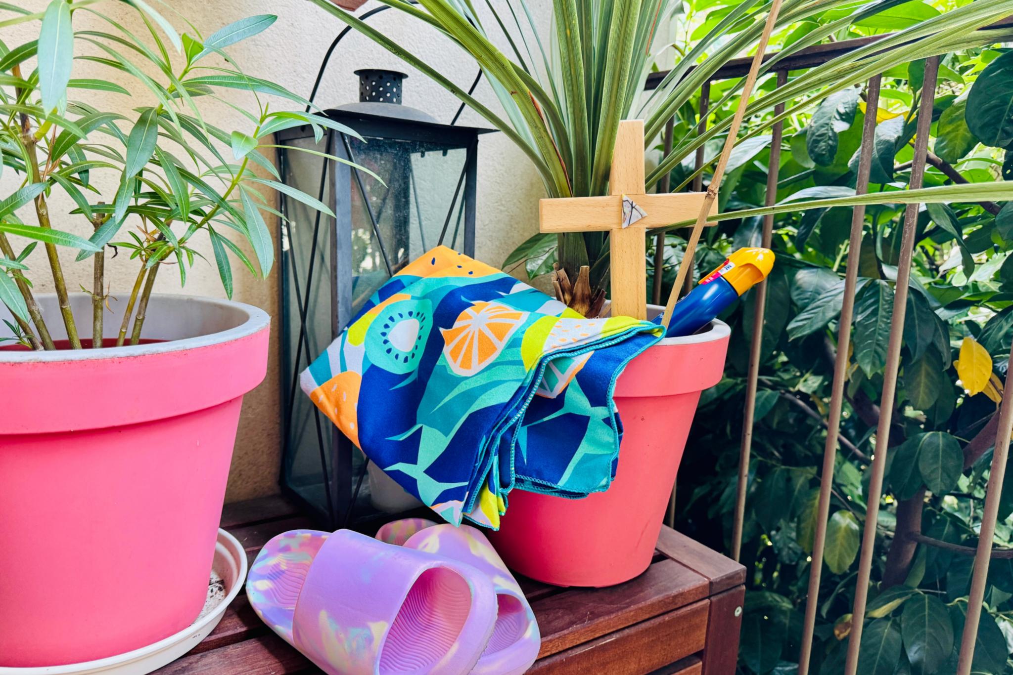 Zwischen Balkonpflanzen steht ein Kreuz, eine Flasche Sonnenmilch, Badeschuhe und ein Badetuch