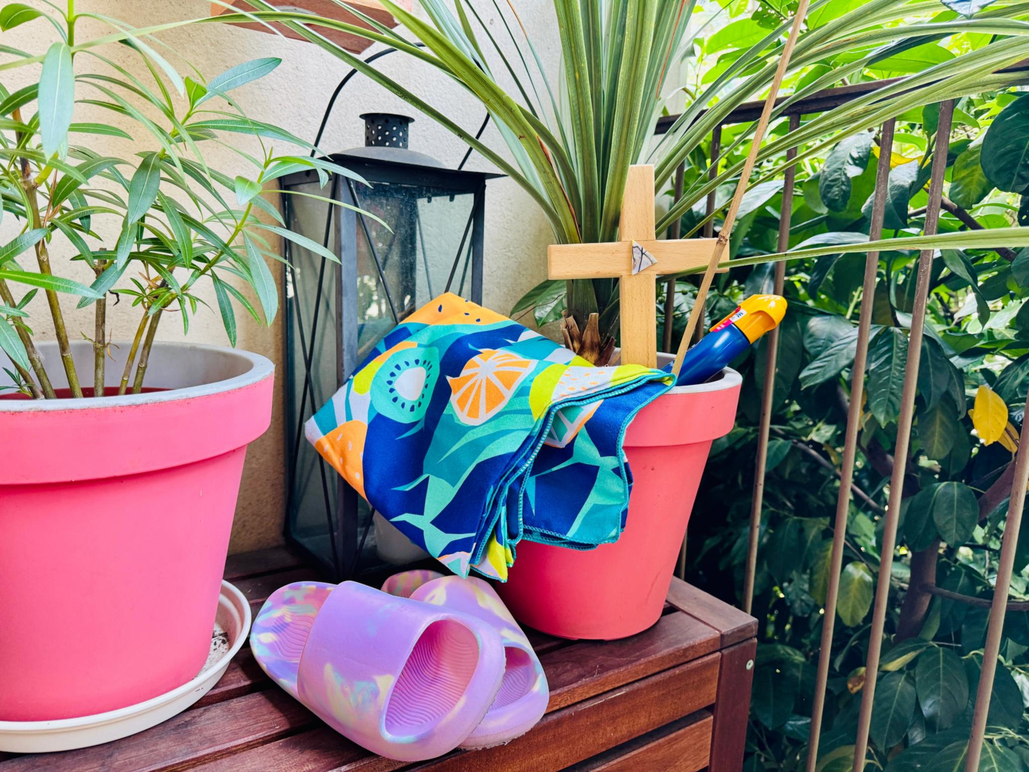 Zwischen Balkonpflanzen steht ein Kreuz, eine Flasche Sonnenmilch, Badeschuhe und ein Badetuch