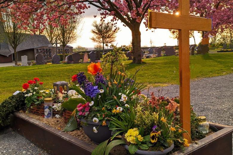 Grab mit Holzkreuz und vielen Frühlingsblumen; im Hintergrund sieht man den friedhof, Wiese und blühende Bäume bei untergehender Sonne