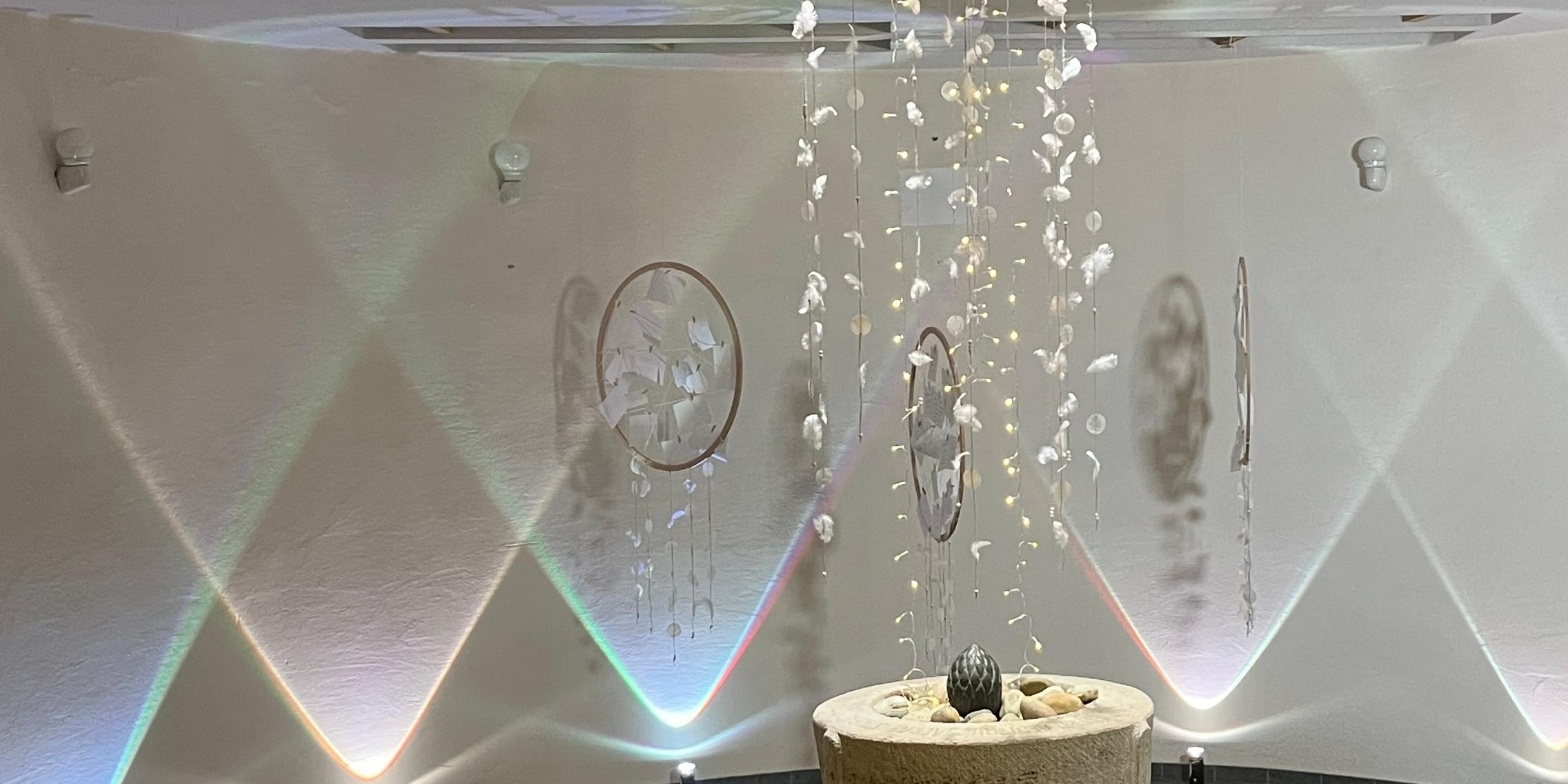 Blick in die Frauenkirche in Köllerbach, hier das umgestaltete ehemalige Taufbecken Wände sind mit geometrischen Mustern bestrahlt, in der Mitte steht ein Taufbecken, darüber hängt ein Windspiel