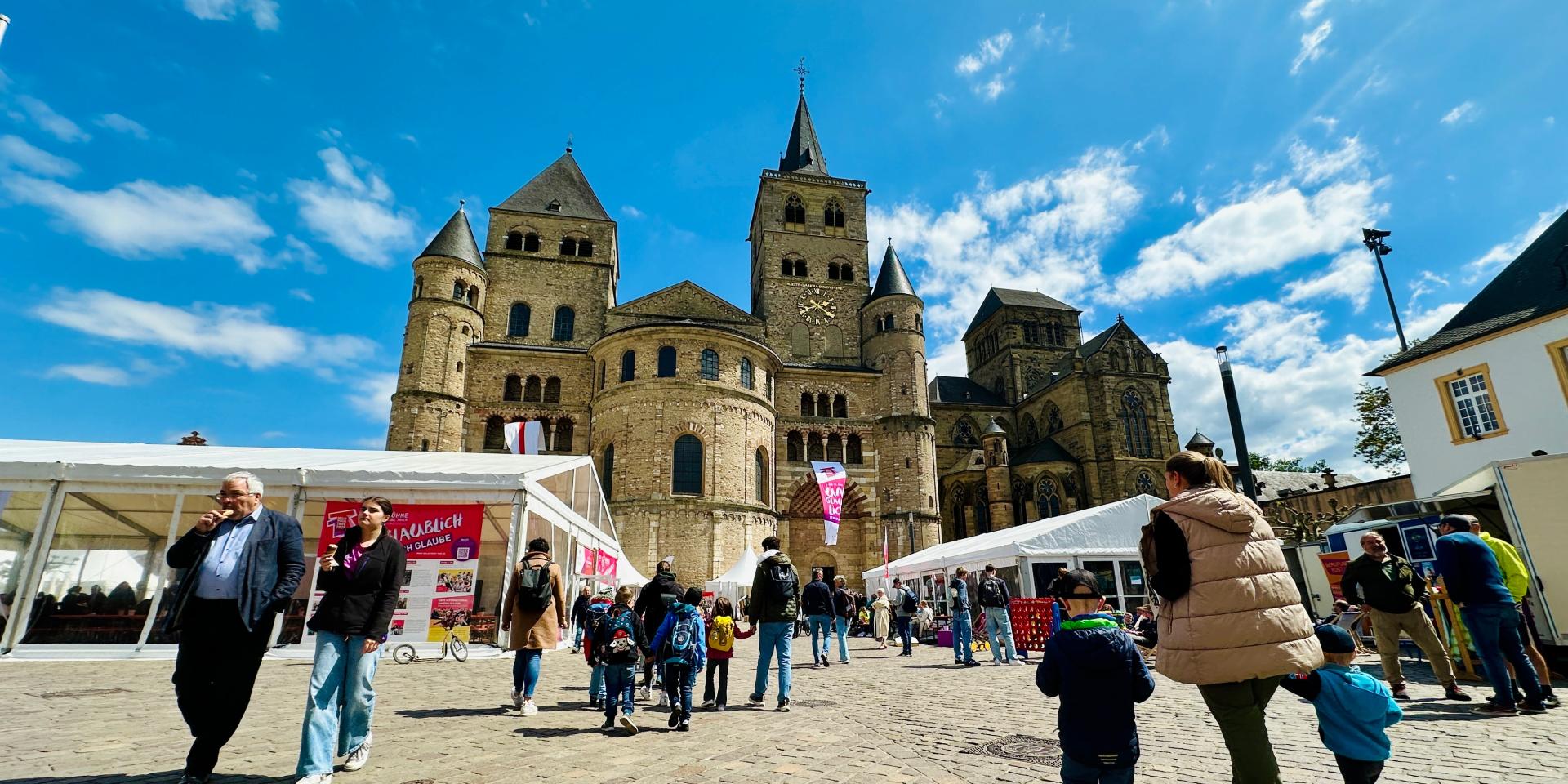 Auf dem Domfreihof tummeln sich mehrere Menschen. Links und rechts sieht man die Festivalzelte, im Hintergrund den Dom vor blauem Himmel.