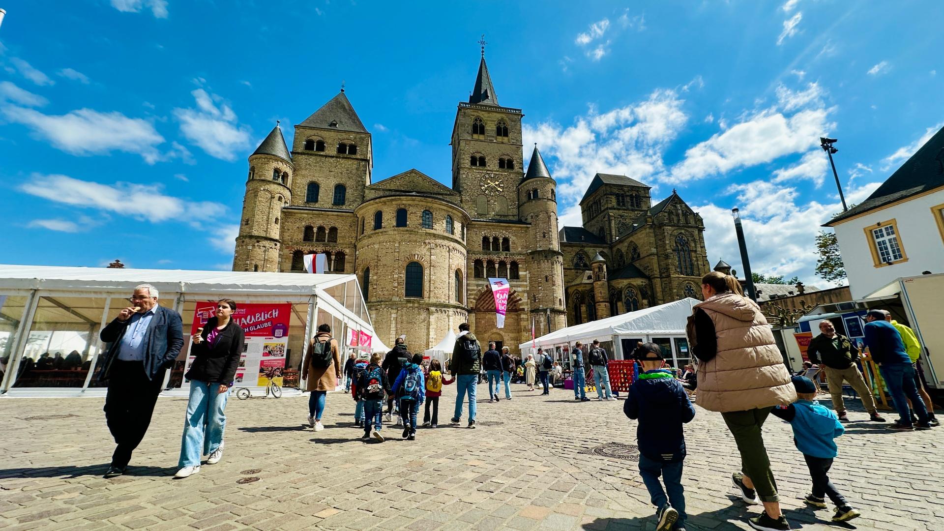 Auf dem Domfreihof tummeln sich mehrere Menschen. Links und rechts sieht man die Festivalzelte, im Hintergrund den Dom vor blauem Himmel.