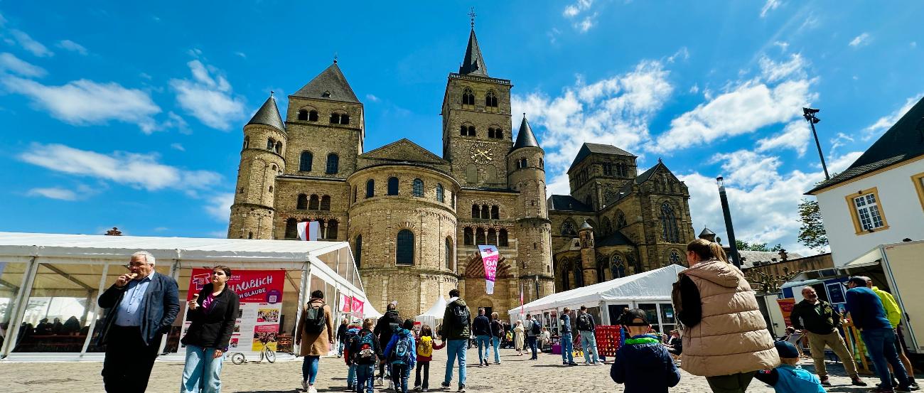 Auf dem Domfreihof tummeln sich mehrere Menschen. Links und rechts sieht man die Festivalzelte, im Hintergrund den Dom vor blauem Himmel.