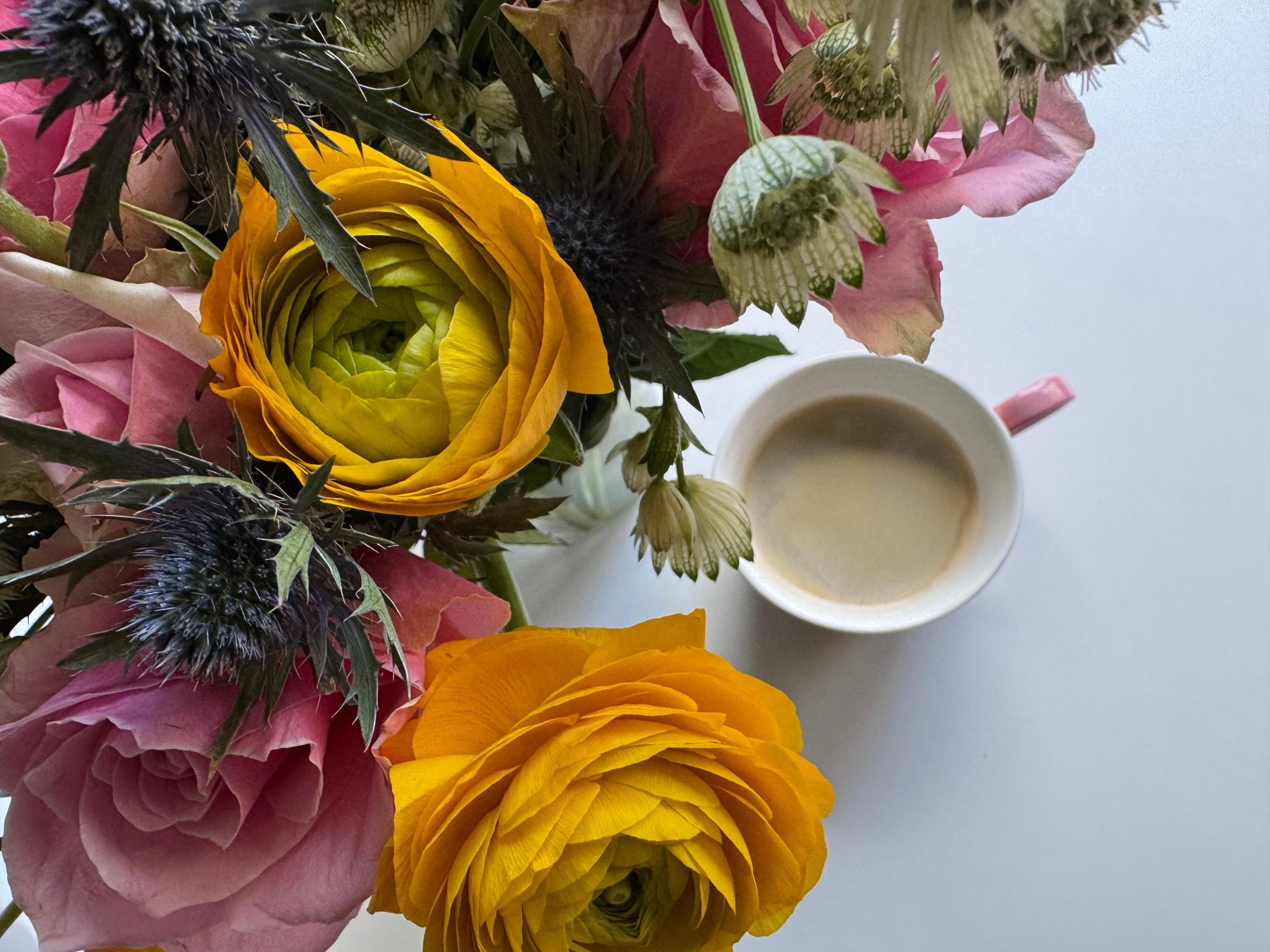 Auf einem Tisch steht eine Tasse Kaffee neben einem Strauß mit Rosen, Ranunkeln und Disteln